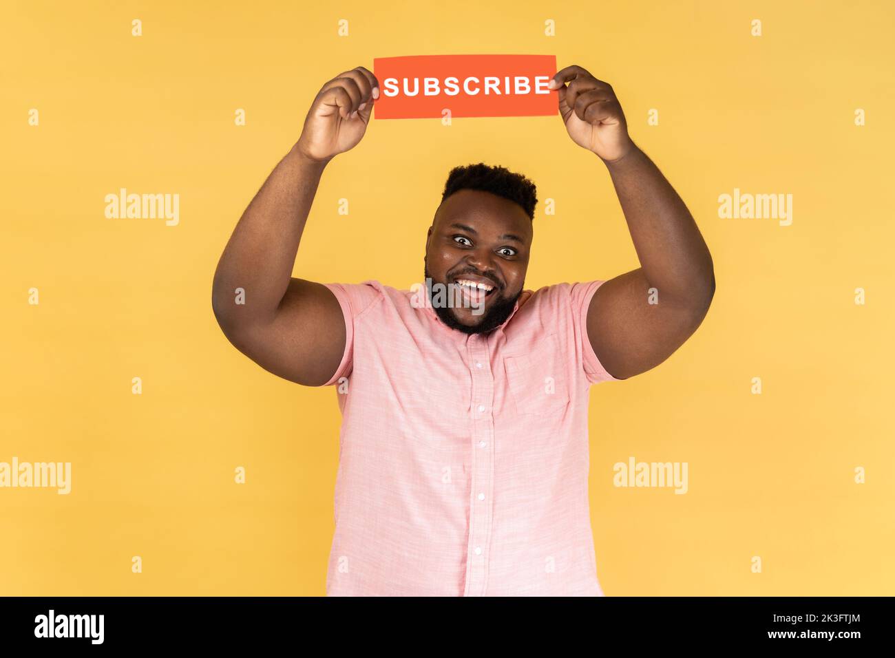 Portrait of excited amazed man in pink shirt holding with paper card with subscribe inscription above head, looking at camera with toothy smile. Indoor studio shot isolated on yellow background. Stock Photo