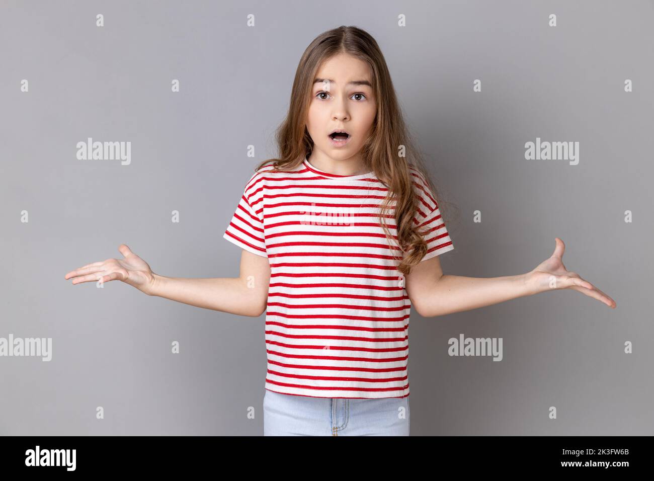 What do you want. Portrait of little girl wearing striped T-shirt standing with raised hands and surprised indignant expression, asking what reason. Indoor studio shot isolated on gray background. Stock Photo