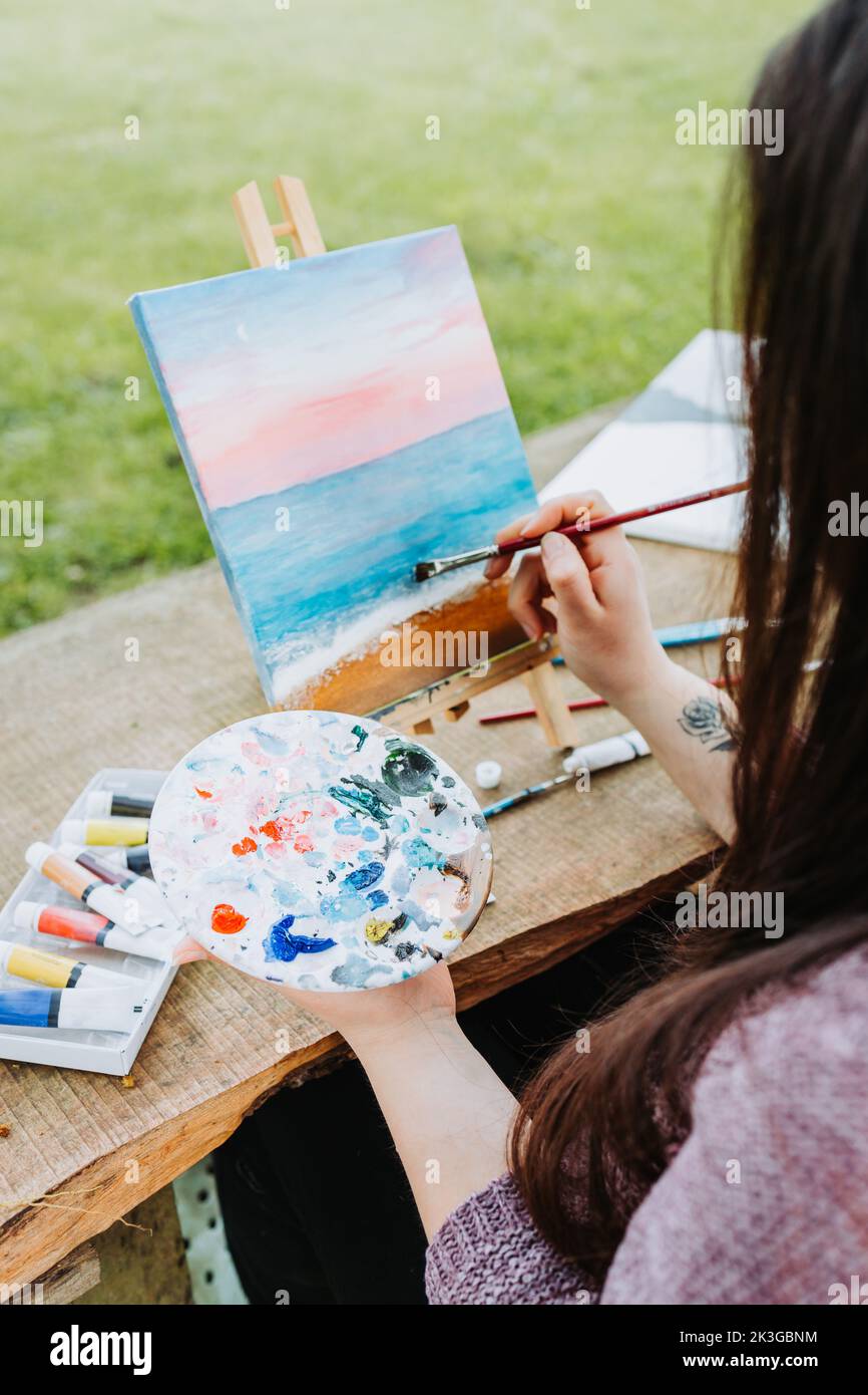 Unrecognizable young female artist painting her own beach scenery with oil on a canvas outdoor. Amateur painter. Stock Photo