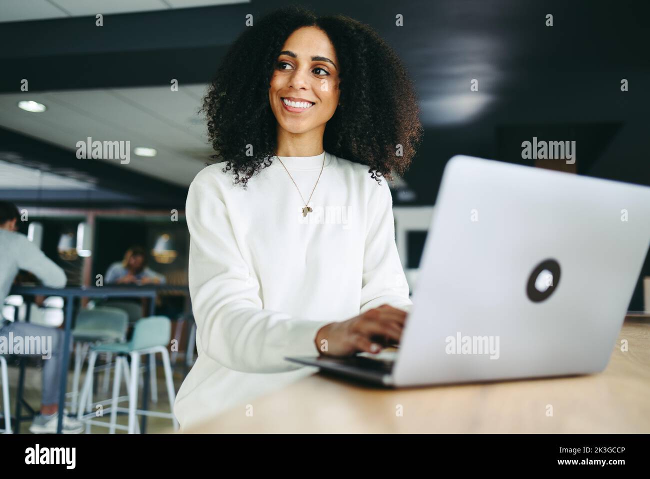 Pensive businesswoman looking away with a smile while typing on a laptop. Happy young businesswoman composing an email while working remotely in a co- Stock Photo