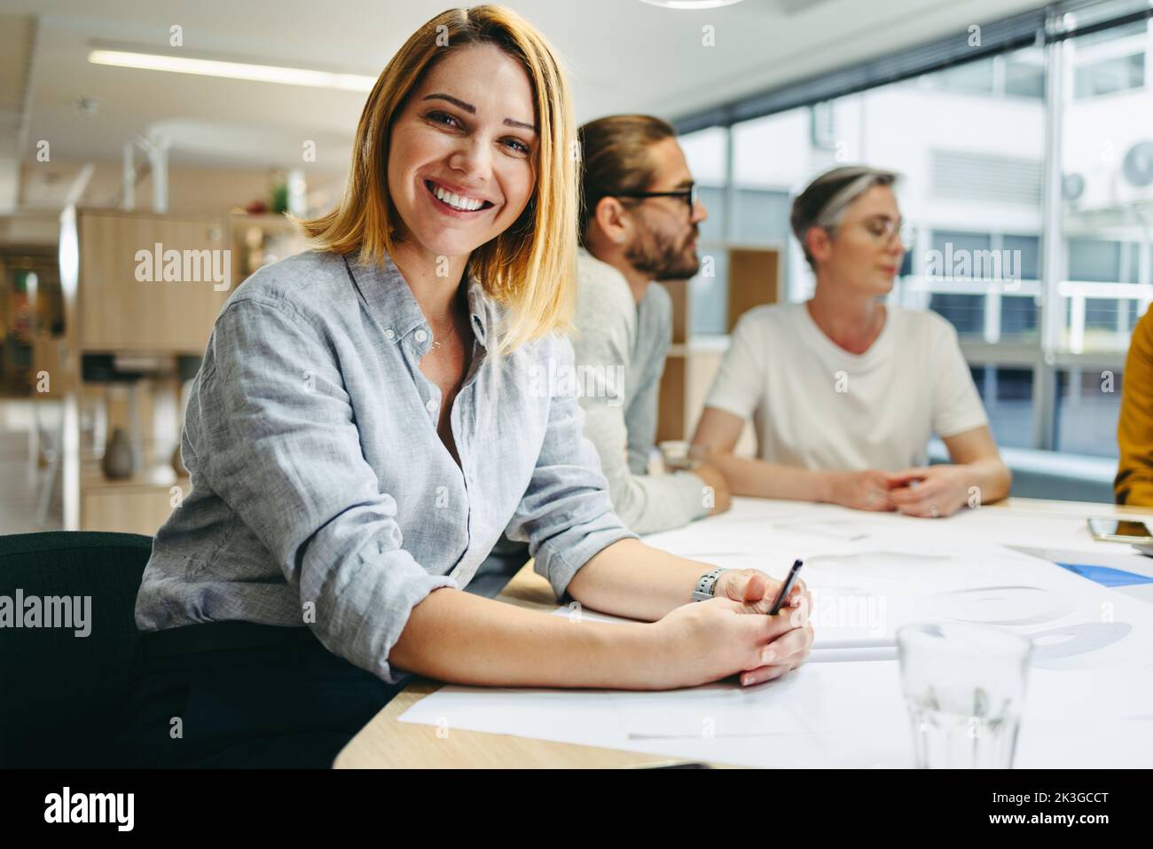 Cheerful young designer smiling at the camera while sitting in a meeting with her colleagues. Team of innovative businesspeople sharing creative ideas Stock Photo