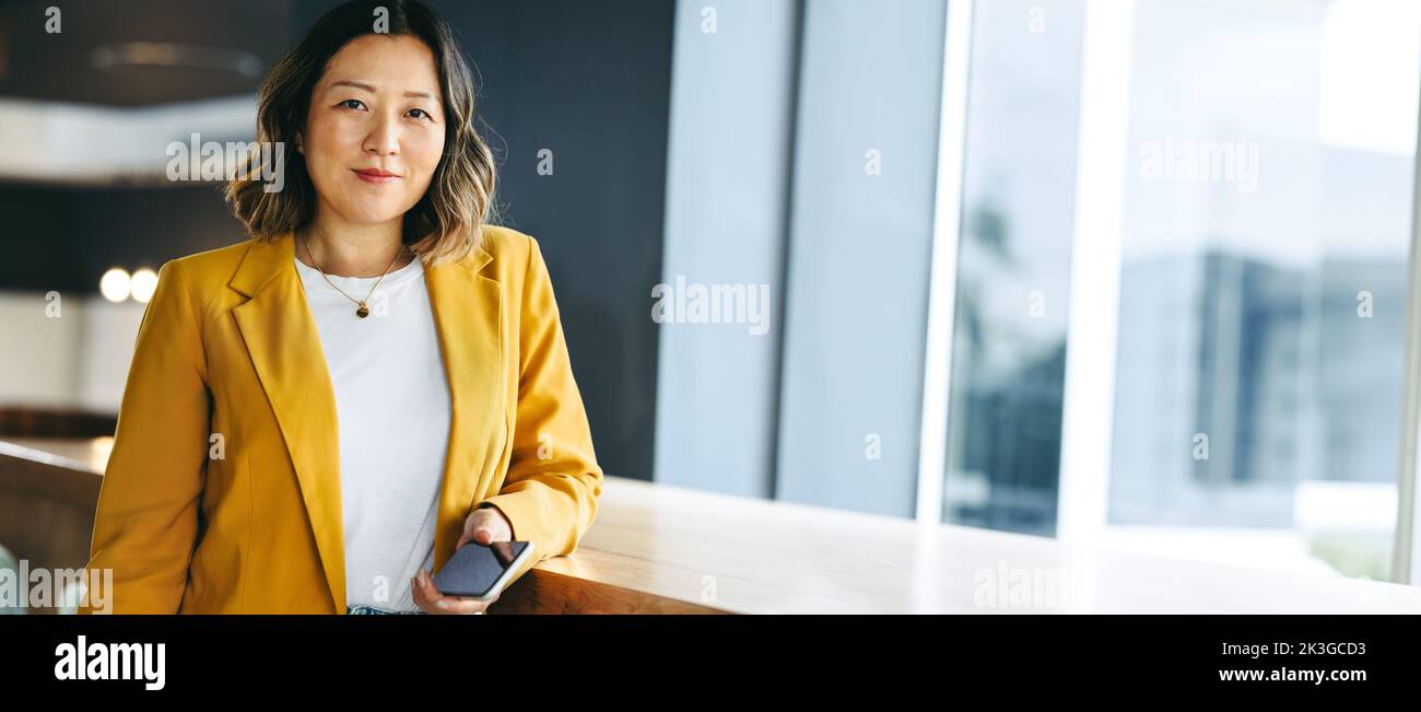 Female entrepreneur looking at the camera while standing with a smartphone in her hand. Ethnic businesswoman working remotely in a co-working space. Stock Photo