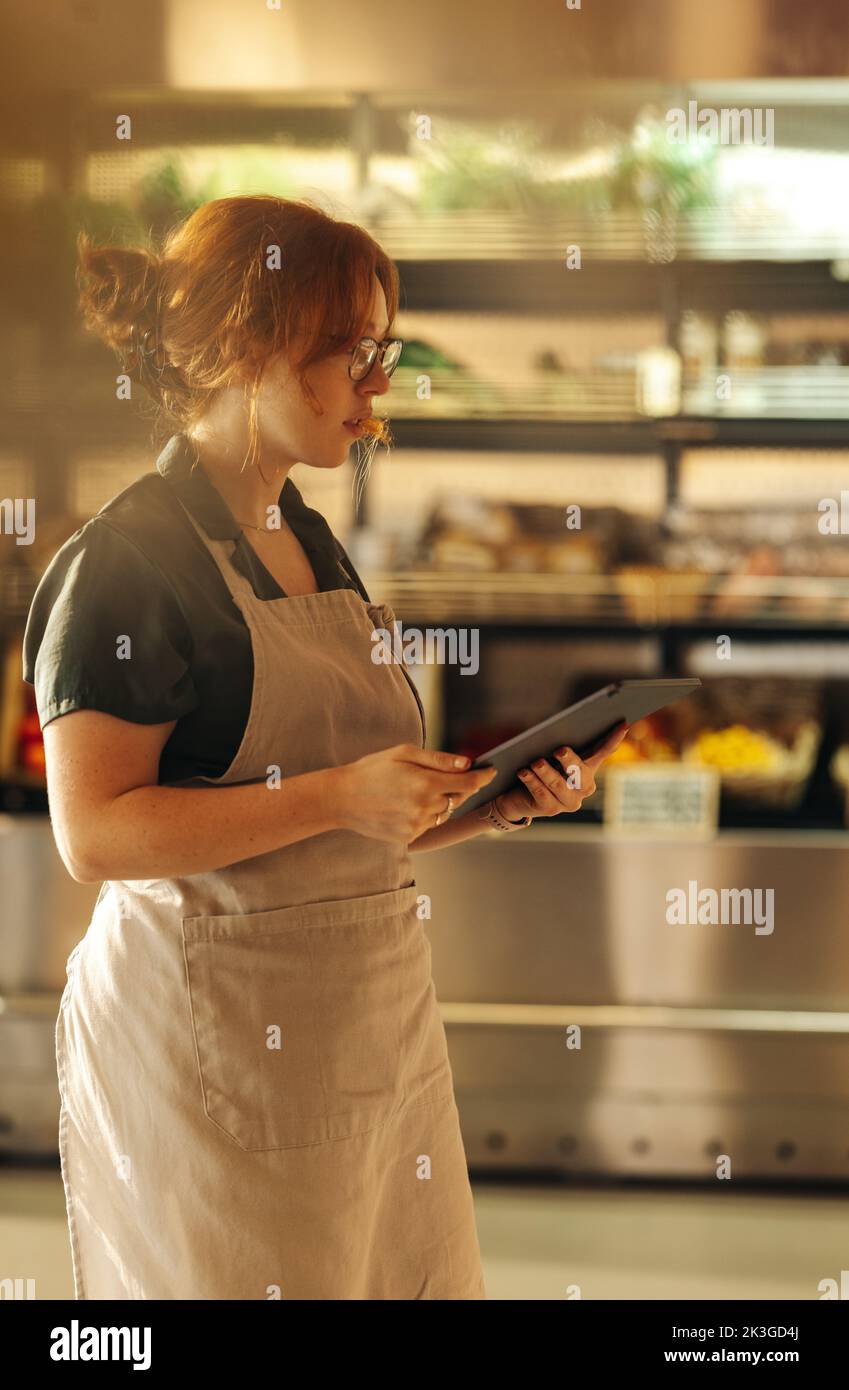 Young grocery store owner taking inventory using a digital tablet in her shop. Successful female entrepreneur running her small business using wireles Stock Photo