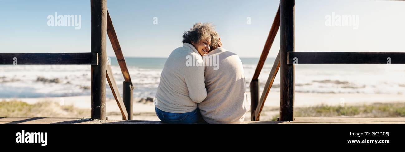 Panoramic view of a happy elderly woman smiling cheerfully while sitting in front of the ocean with her husband. Retired senior couple spending some q Stock Photo