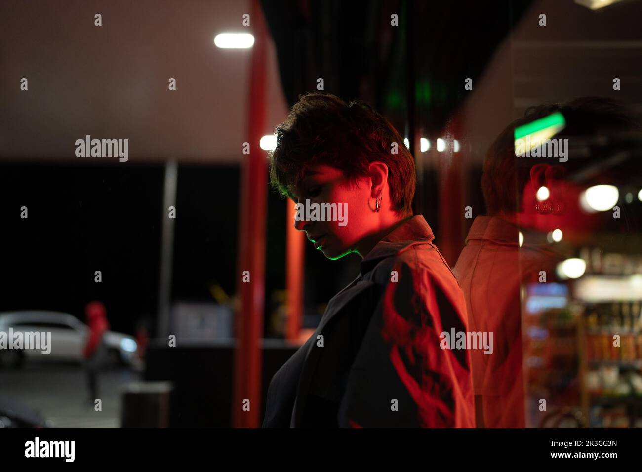 Pensive woman against glass wall on the street with red neon light at night. Sad and loneliness. Stock Photo