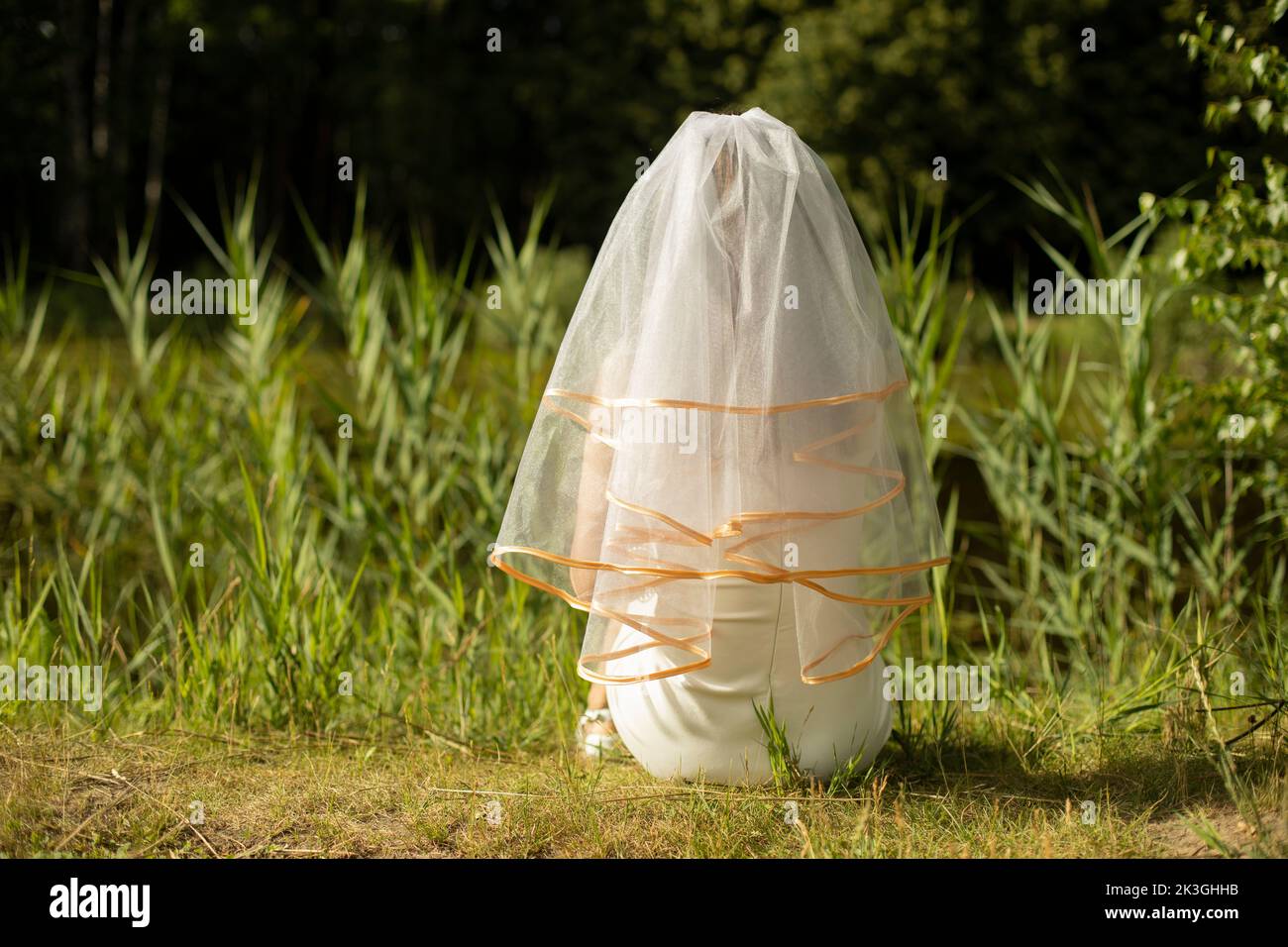 Bride on shore of lake. Girl in white dress in park. Fabric on head. Woman waiting for wedding. Summer day and white clothes. Stock Photo