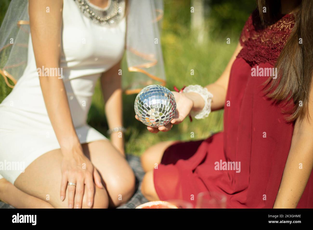 Girls in summer in dresses sit on grass. Girl holds ball in her hand. Mirror ball in palm of your hand. Details of outdoor recreation. Stock Photo