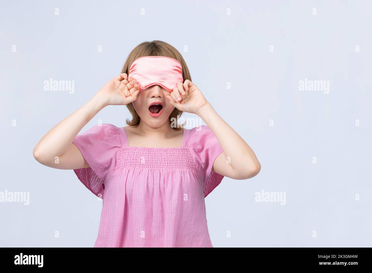 Sleepy tired lovely asian teen girl covering mouth with hand and yawning wearing sleep mask on white background Stock Photo