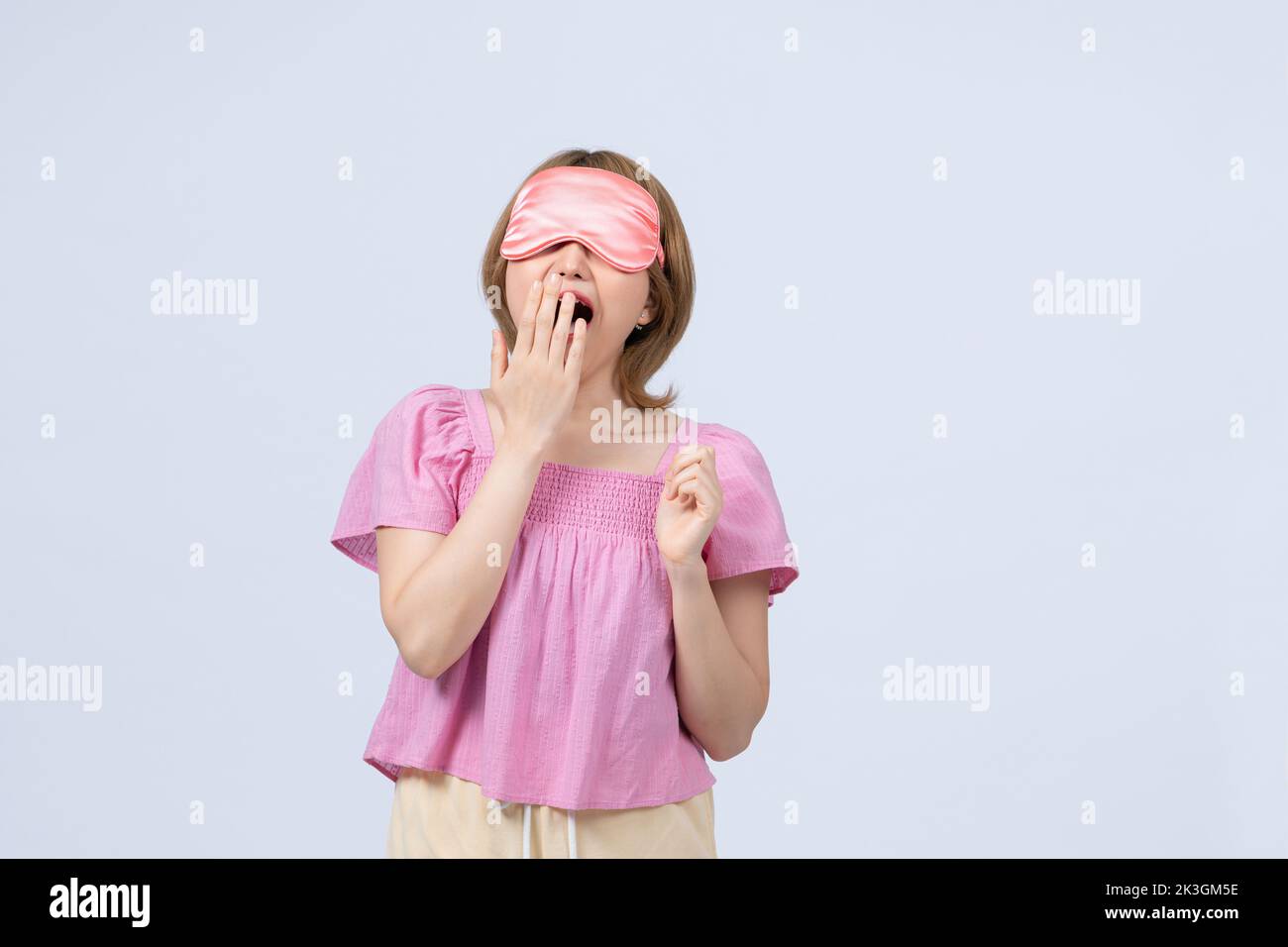 Tired sleepy young asian female yawns, covers mouth with palm, wears sleep mask on head, Stock Photo