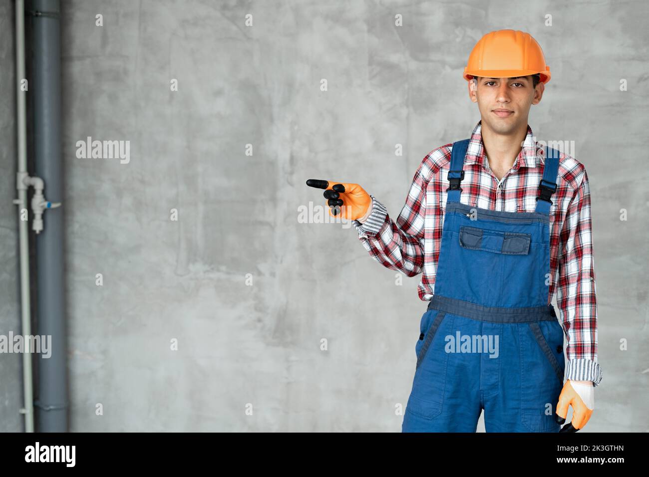 Young muslim man builder over grey concrete wall wearing contractor uniform and safety helmet showing with thumb up to the side with happy face smilin Stock Photo