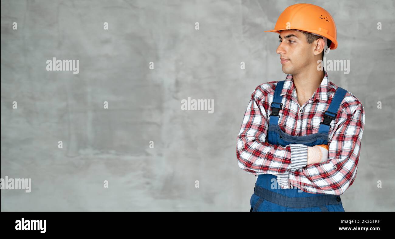 Portrait of confident arabic builder in overall and shirt having arms crossed, looking at side, isolated on gray wall background, indoors. house renov Stock Photo