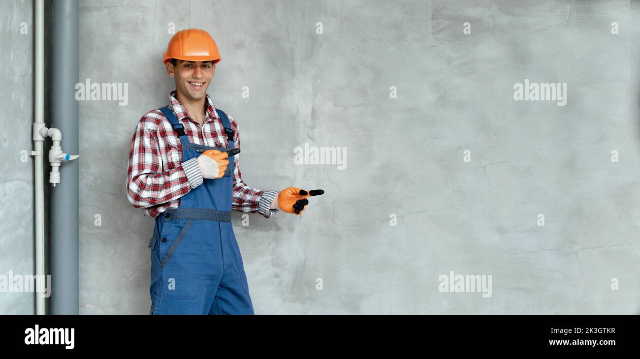 Young muslim man over grey wall wearing overalls and safety helmet pointing and showing with thumb up to the side with happy face smiling. Banner, spa Stock Photo