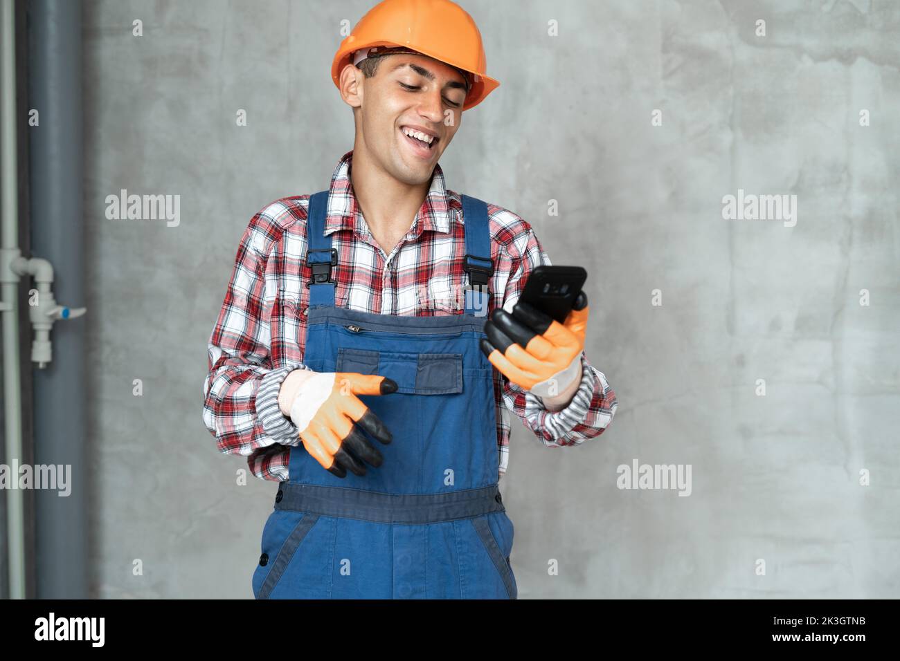 happy smiling male worker or builder in yellow helmet and overalls with smartphone over gray wall background. profession, construction and building co Stock Photo