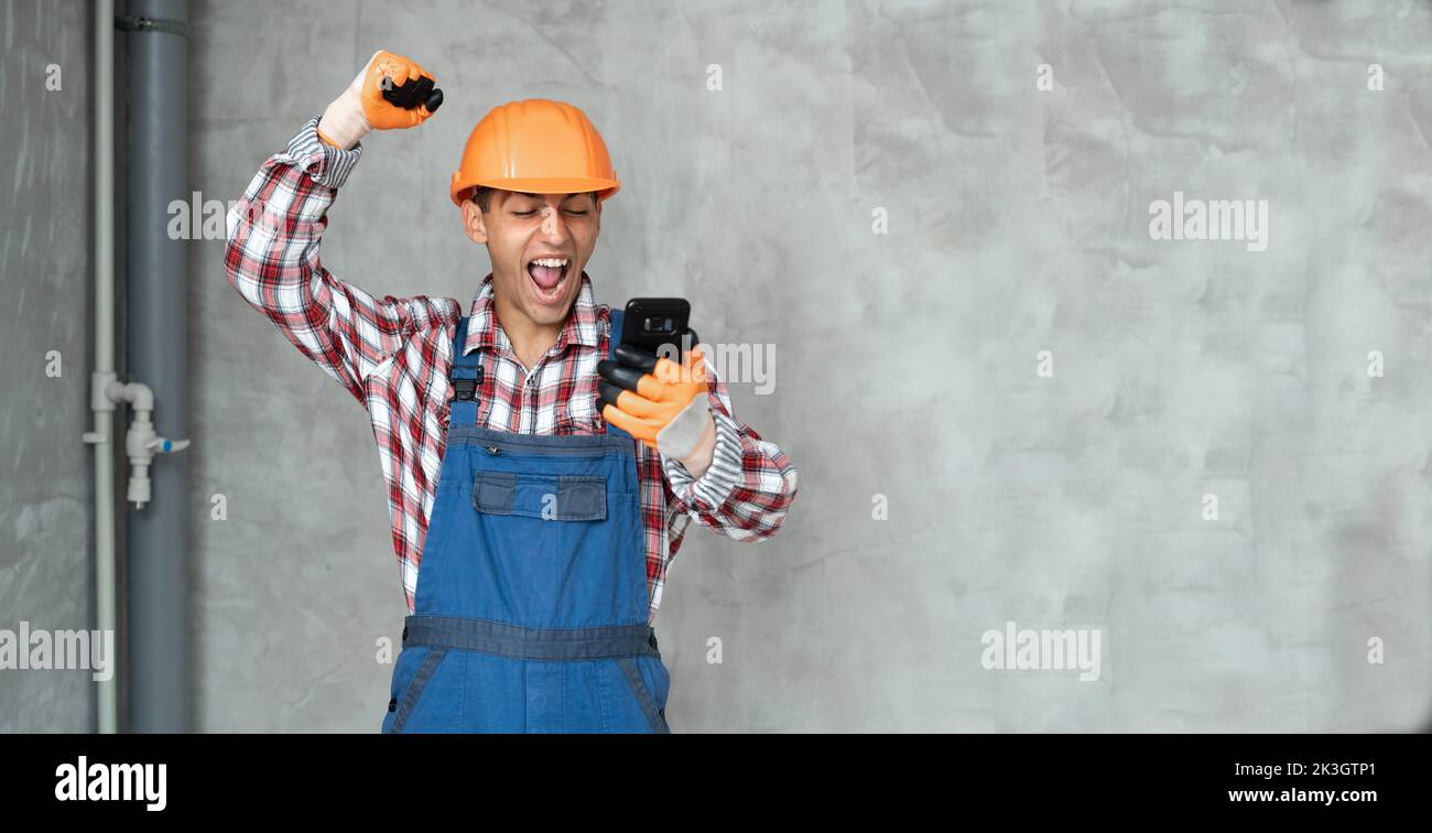 Photo of a happy positive young man builder in helmet posing isolated over gray wall background using mobile phone. Human positive emotion concept. ba Stock Photo