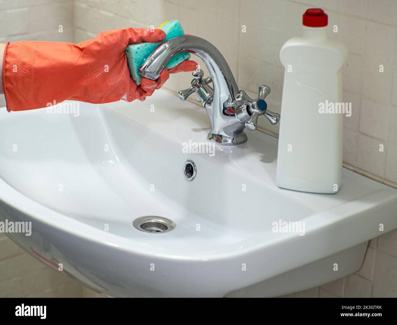 Cleaning bathroom sink and faucet with detergent in orange rubber gloves with green sponge