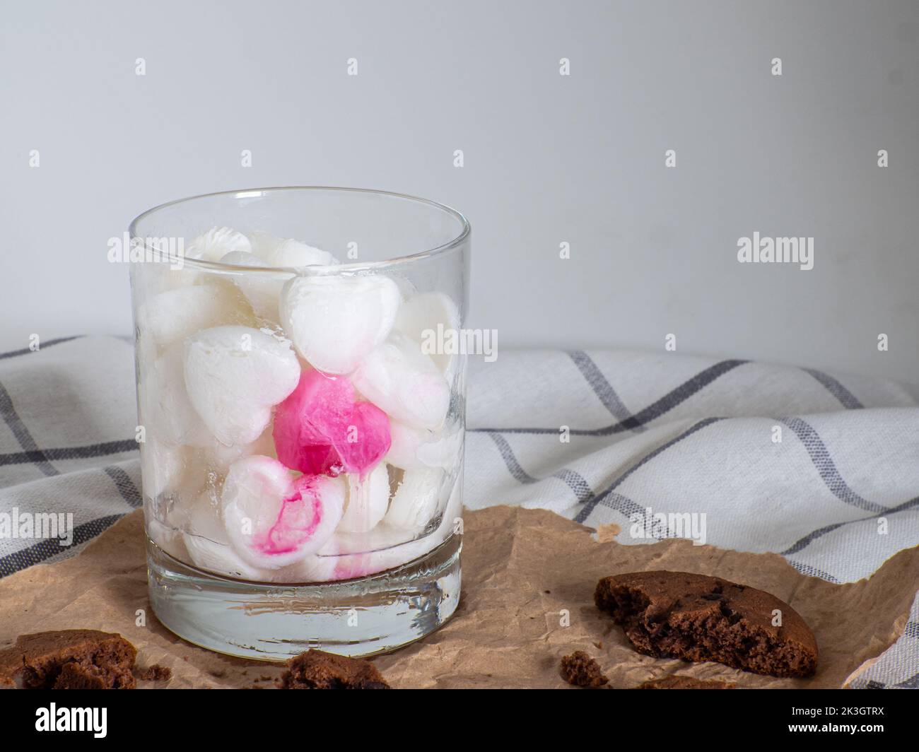 White Ice cubes made from milk in heart shape with one red heart in glass. Space for text. Cocoa cookies with chocolate on paper on a white background Stock Photo