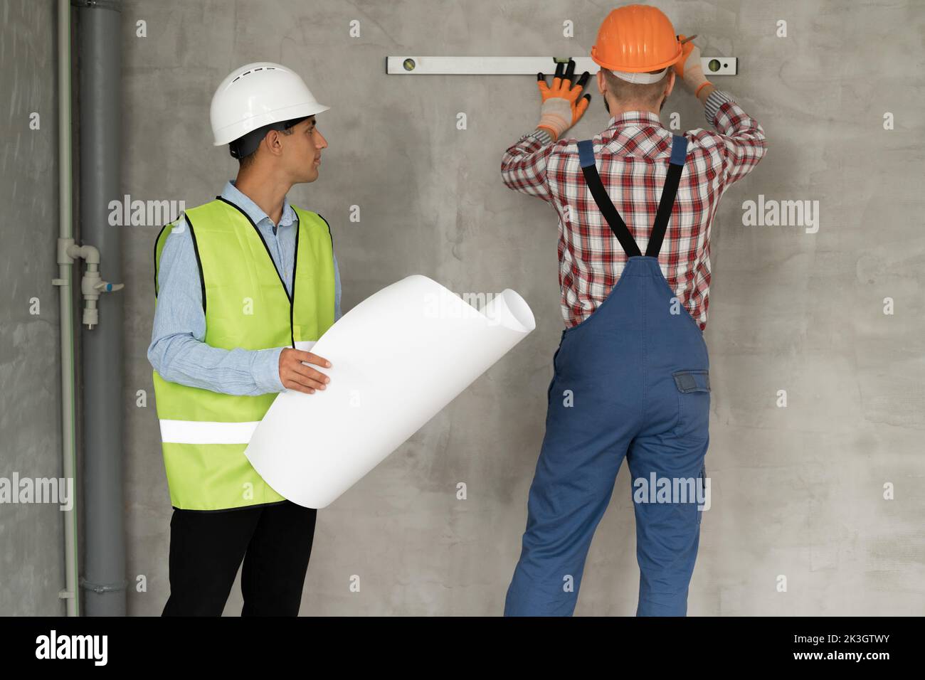 A young civil engineer with blueprints in his hands checks the work of a builder doing repairs in the house. Builder in a protective helmet measures t Stock Photo