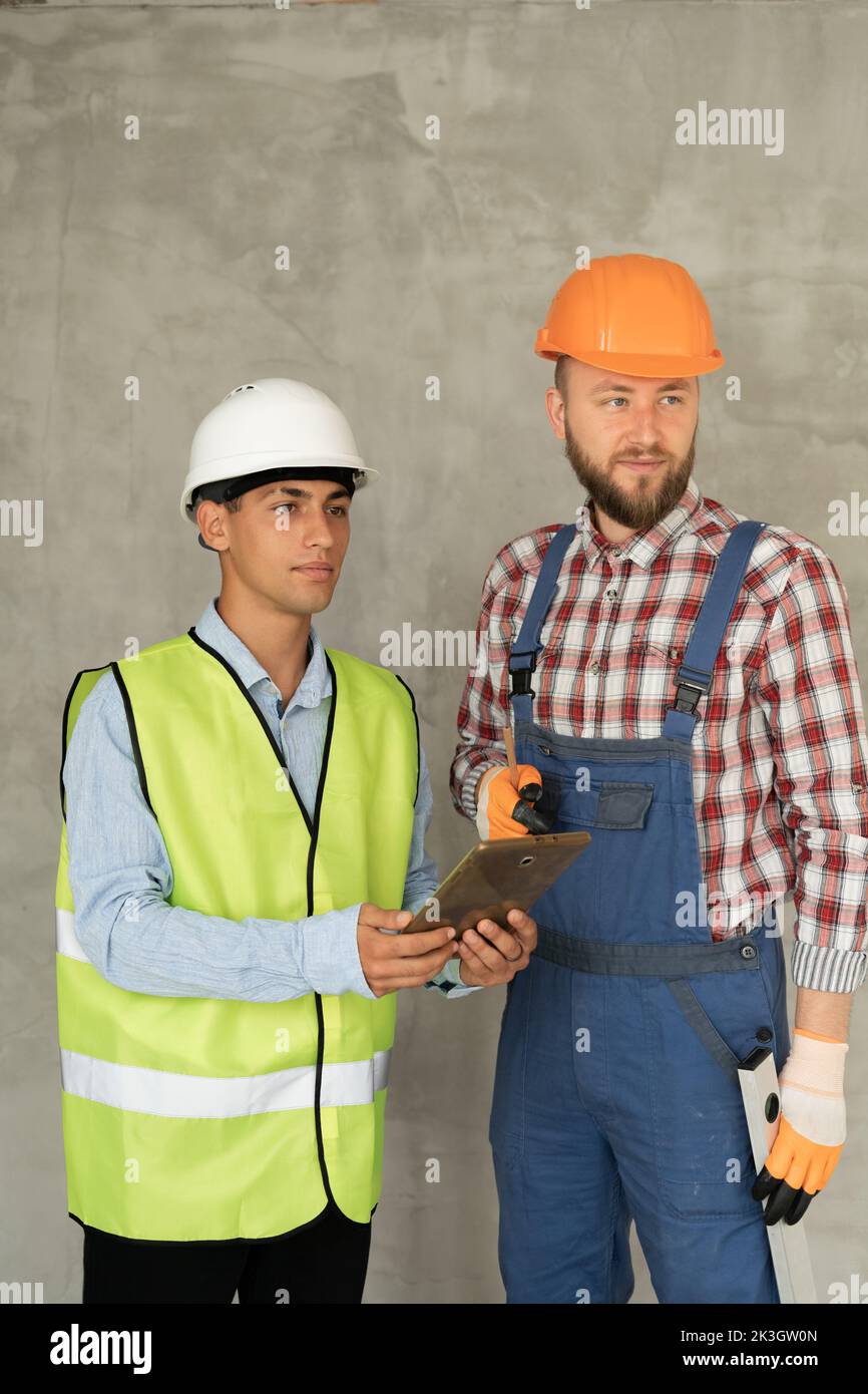 Working in team. Two young and cheerful builders in protective helmets are using digital tablet and working while standing on gray background. Copy sp Stock Photo