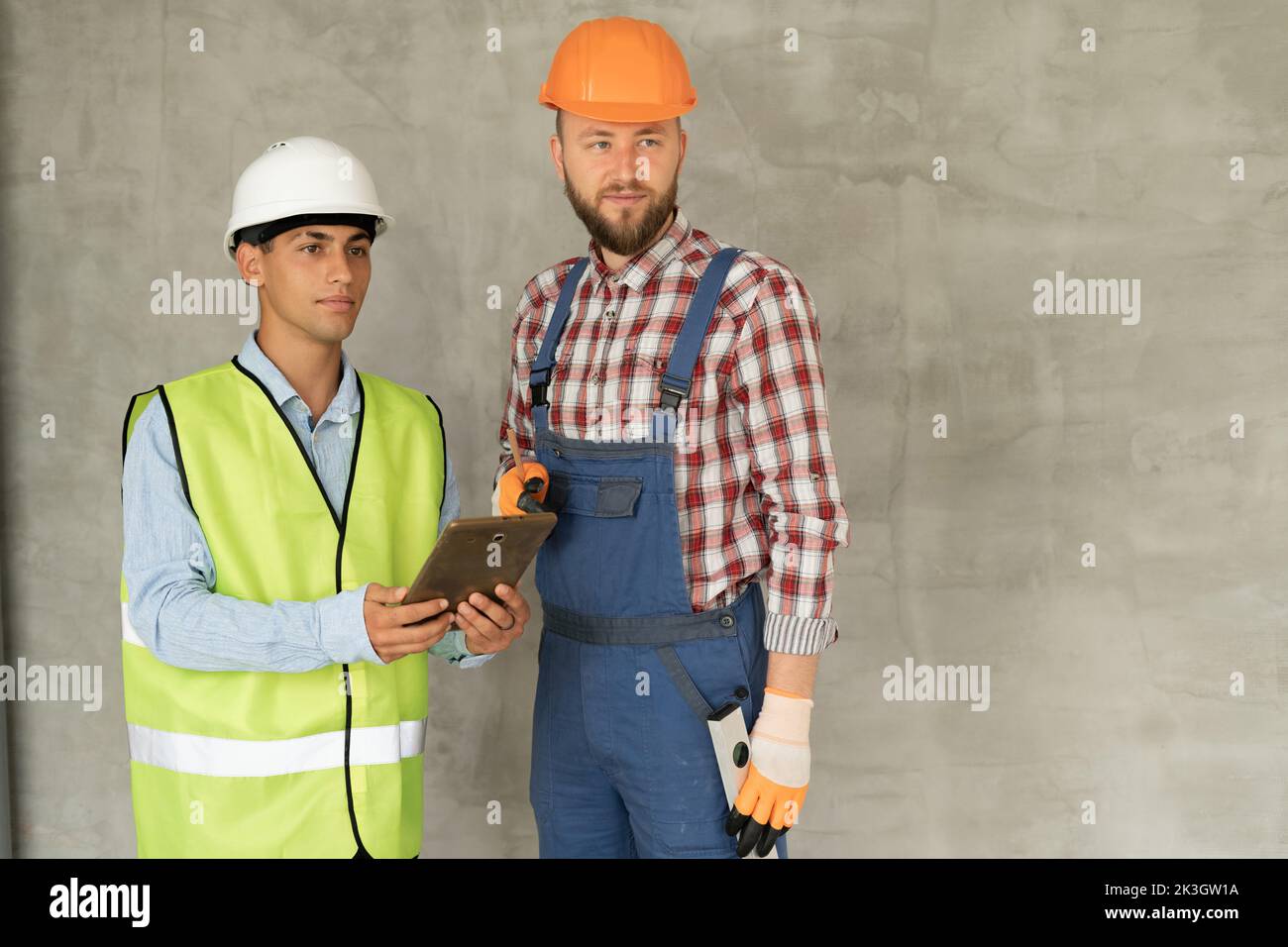 Construction worker and engineer on site discussing blueprints on pad or tablet computer, gray wall background. Place for text Stock Photo