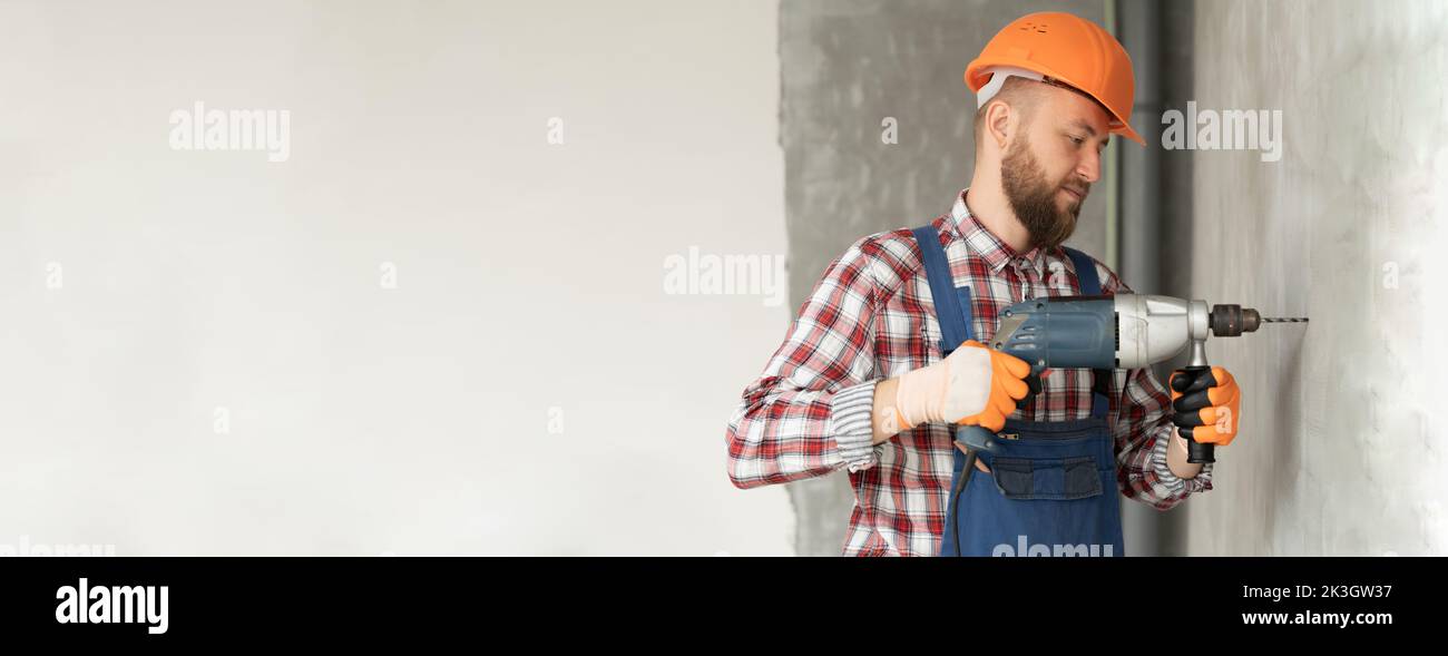 Laborer man in protective gloves drills concrete wall with drill. Repair service and construction concept. Banner. Space for text Stock Photo