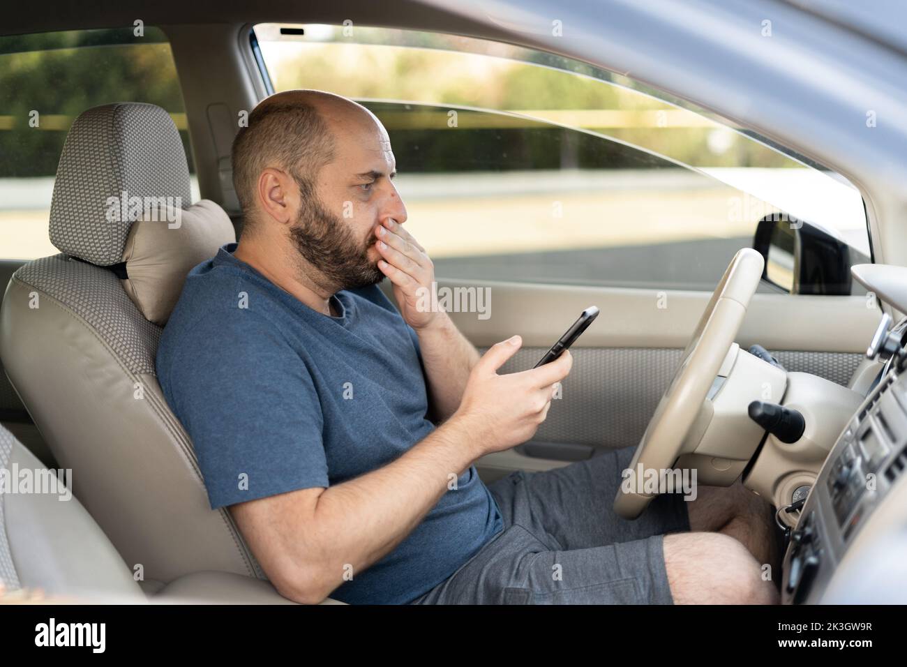 Man sitting inside car with mobile phone in hand texting. Shocked man checking his smartphone not paying attention at road stunned by bad text message Stock Photo