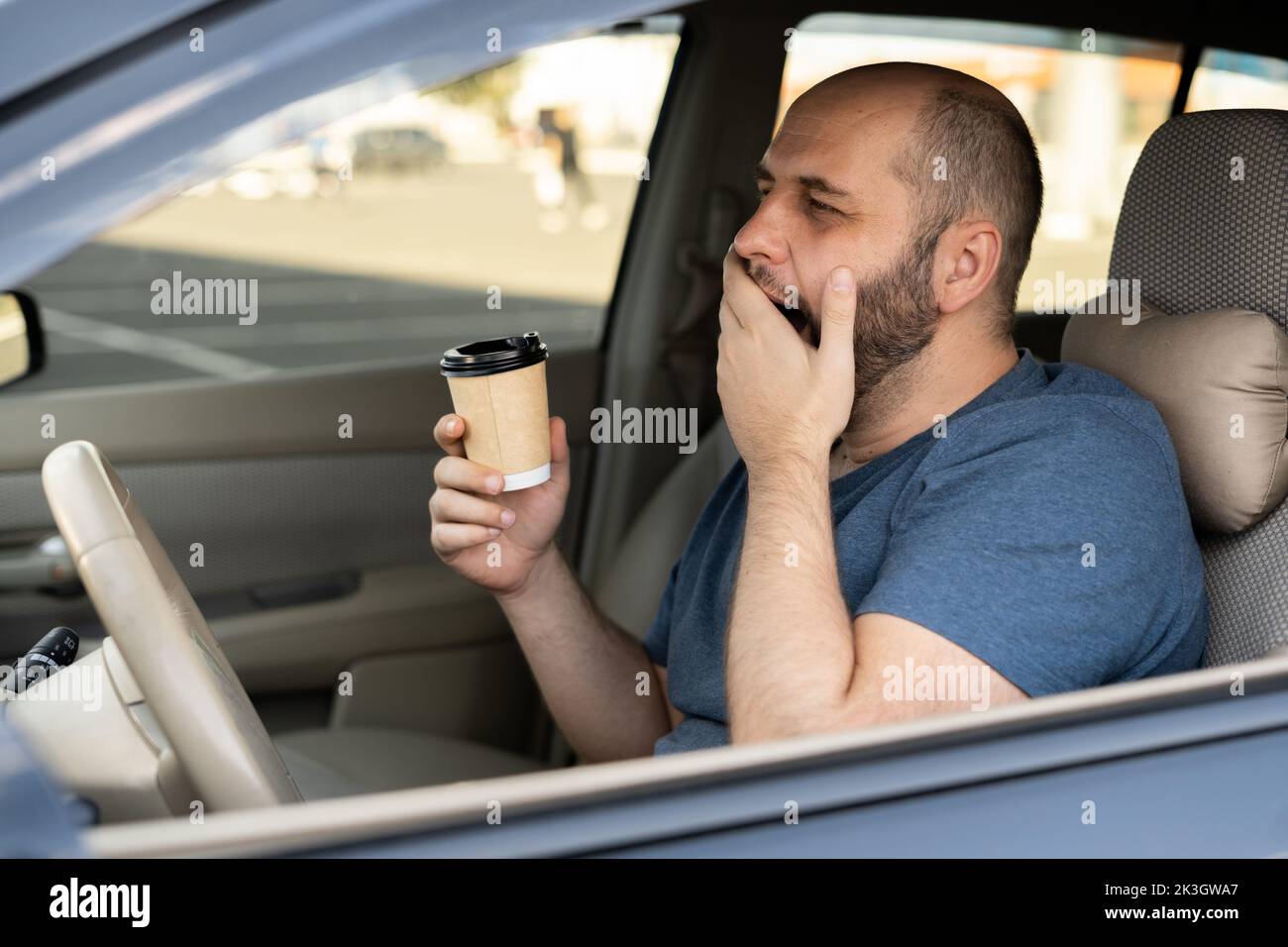 Adult handsome man feeling tired and yawning while driving a car. Driver having coffee early morning inside avto Stock Photo