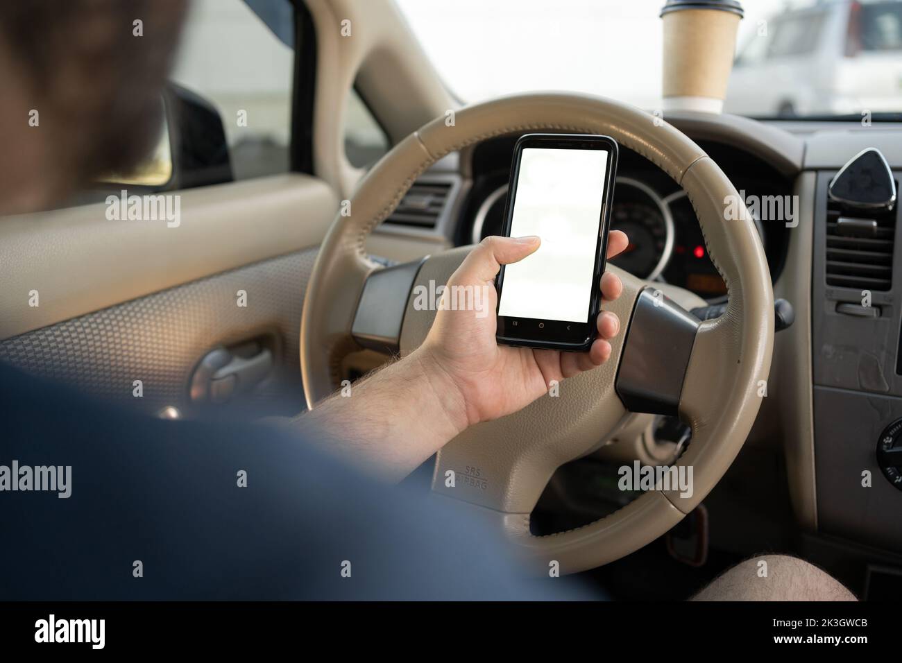 Close-up of male hand holding smartphone with white mockup on screen, background of car steering wheel, searching location via gps navigator applicati Stock Photo