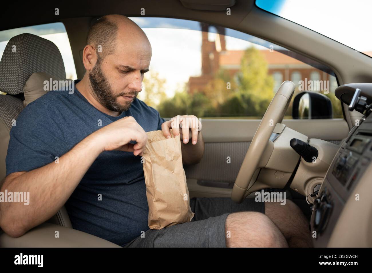 The driver of the car unpacks a package with a hamburger while standing in a traffic jam on the road. Fast food and takeaway concept Stock Photo