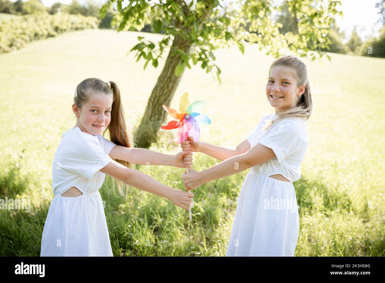 two pretty girls, sisters with white dresses holding colorful pinwheels and standing in green meadow in front of a walnut tree and are happy Stock Photo