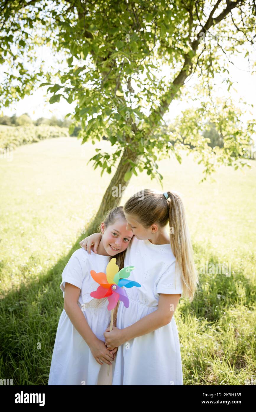 two pretty girls, sisters with white dresses holding colorful pinwheels and standing in green meadow in front of a walnut tree and are happy Stock Photo