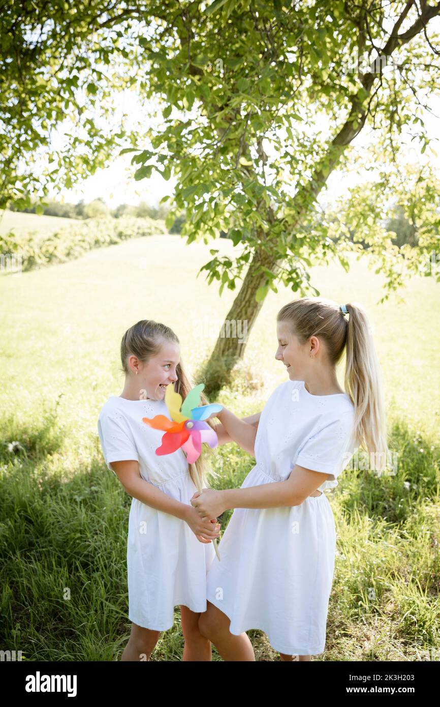 two pretty girls, sisters with white dresses holding colorful pinwheels and standing in green meadow in front of a walnut tree and are happy Stock Photo