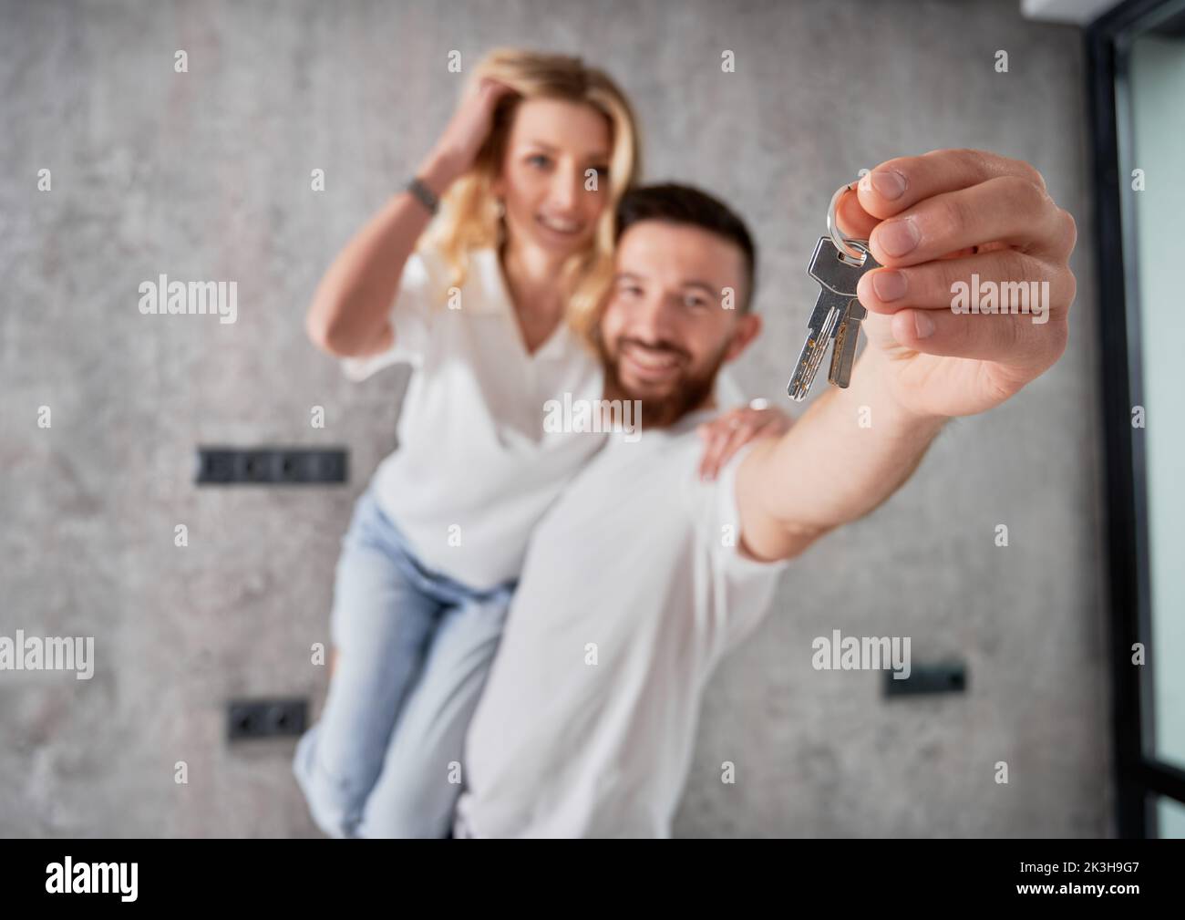 Cheerful man holding wife and smiling while showing apartment keys. Happy couple real estate house buyers demonstrating keys from new home. Focus on male hand with keys. Stock Photo