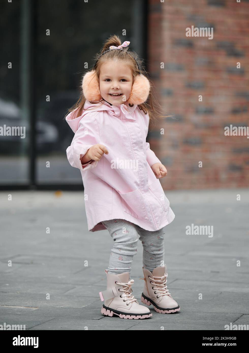 Full length of cute child in pink coat looking at camera and smiling while standing on the street. Happy cheerful kid spending time outdoors in the city. Childhood concept. Stock Photo