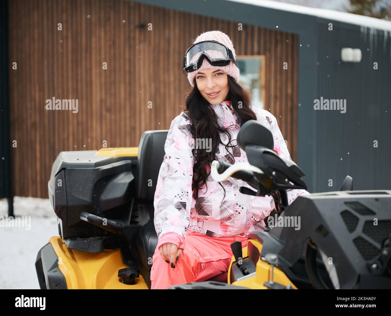 Portrait of beautiful woman posing on offroad four-wheeler ATV with wooden house on background. Concept of active leisure and winter activities. Stock Photo