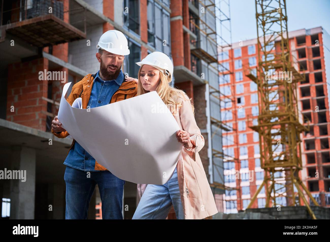 Couple in safety helmets studying architectural drawings outside apartment building under construction. Man and woman discussing building plan outdoors at construction site. Stock Photo