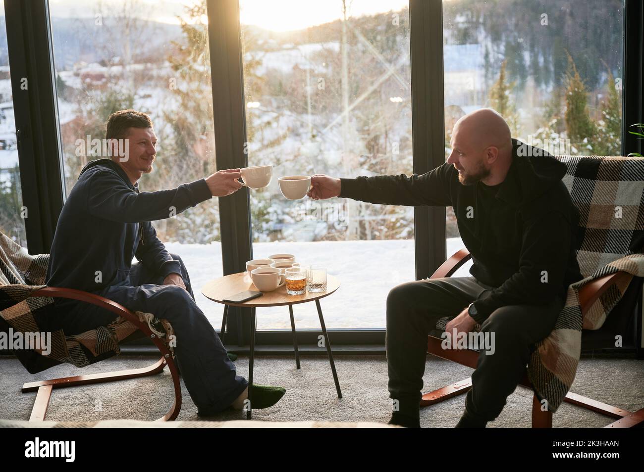 Two young men enjoying view of winter mountain landscape through panoramic windows in contemporary barn house. Happy male tourists sitting on chair, clinking cups of tea in new cottage. Stock Photo