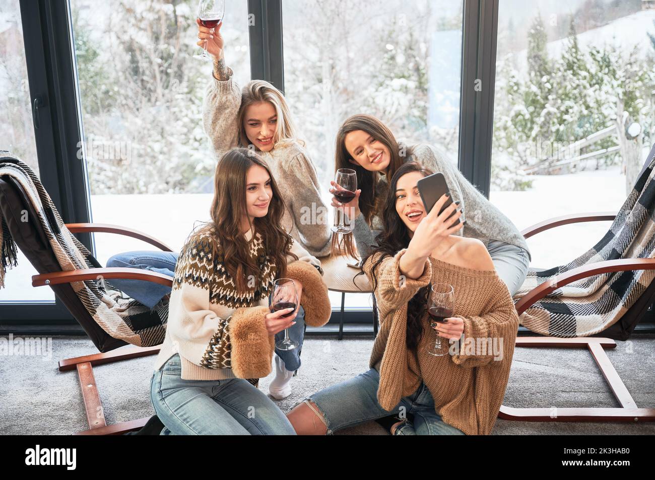 Young women enjoying winter weekends inside contemporary barn house. Four girls having fun, taking selfies on phone and drinking wine. Stock Photo