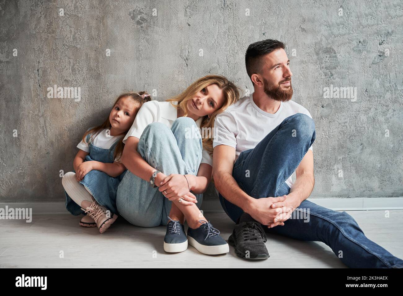 Adorable little girl and parents sitting by the wall in new apartment. Woman leaning head on husband shoulder while resting with family in new home. Stock Photo