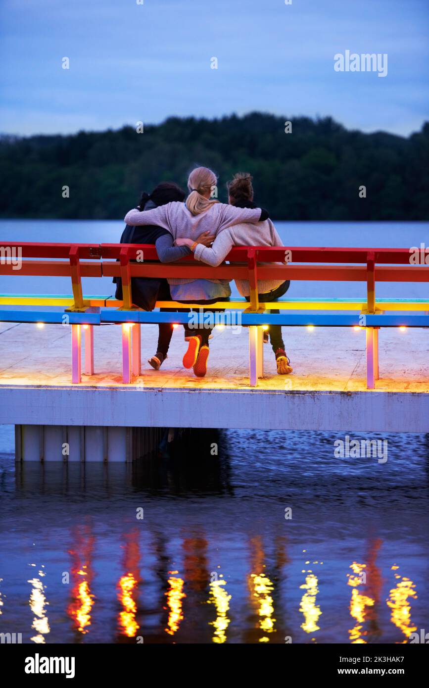 Moments of romance. Rearview shot of three women sitting on a bench on a pier over a lake. Stock Photo