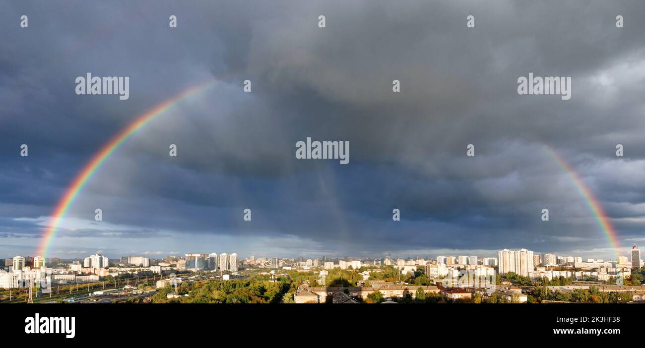 Large rainbow semicircle in the cloudy sky over the city. City landscape after the rain in a light blur. Stock Photo
