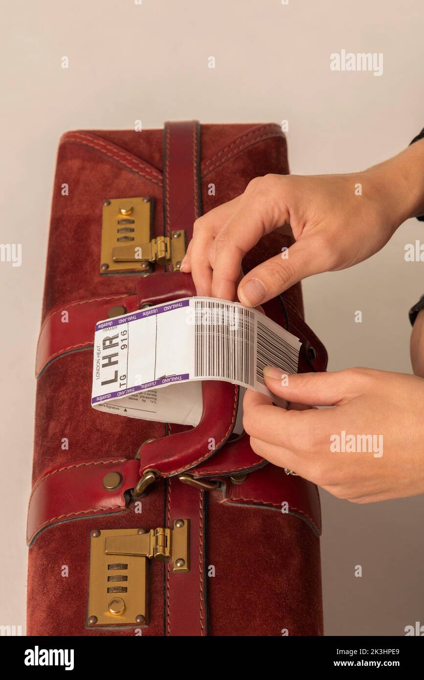 Enngland, UK. 2022. Airline luggage destination identity tag being attached to a dark red leather suitcase handle by womans hands. Stock Photo