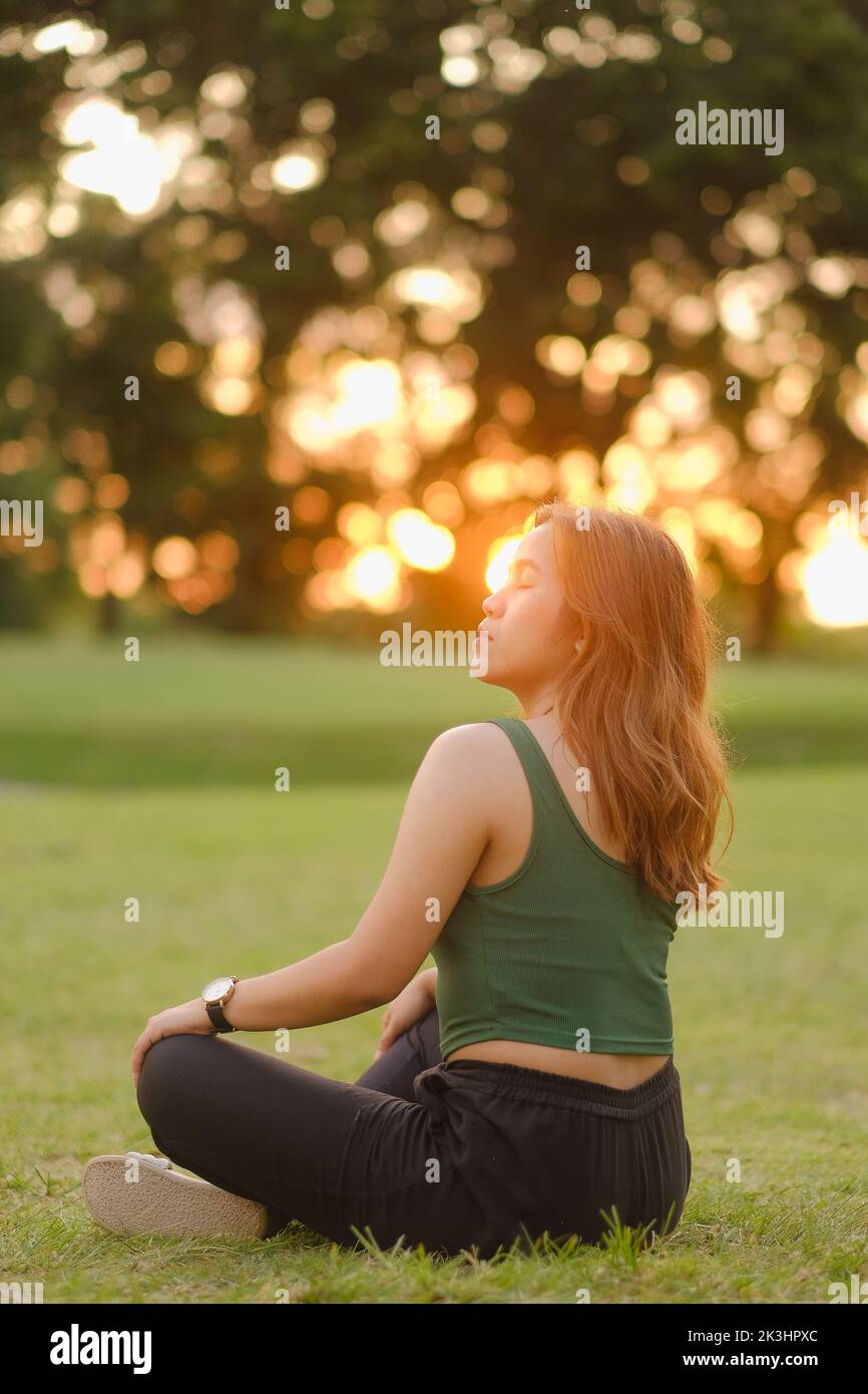 Young Asian woman with long wavy hair, green tank top, and black pants sits cross-legged on the grass and basks in the sunset with eyes closed. Stock Photo
