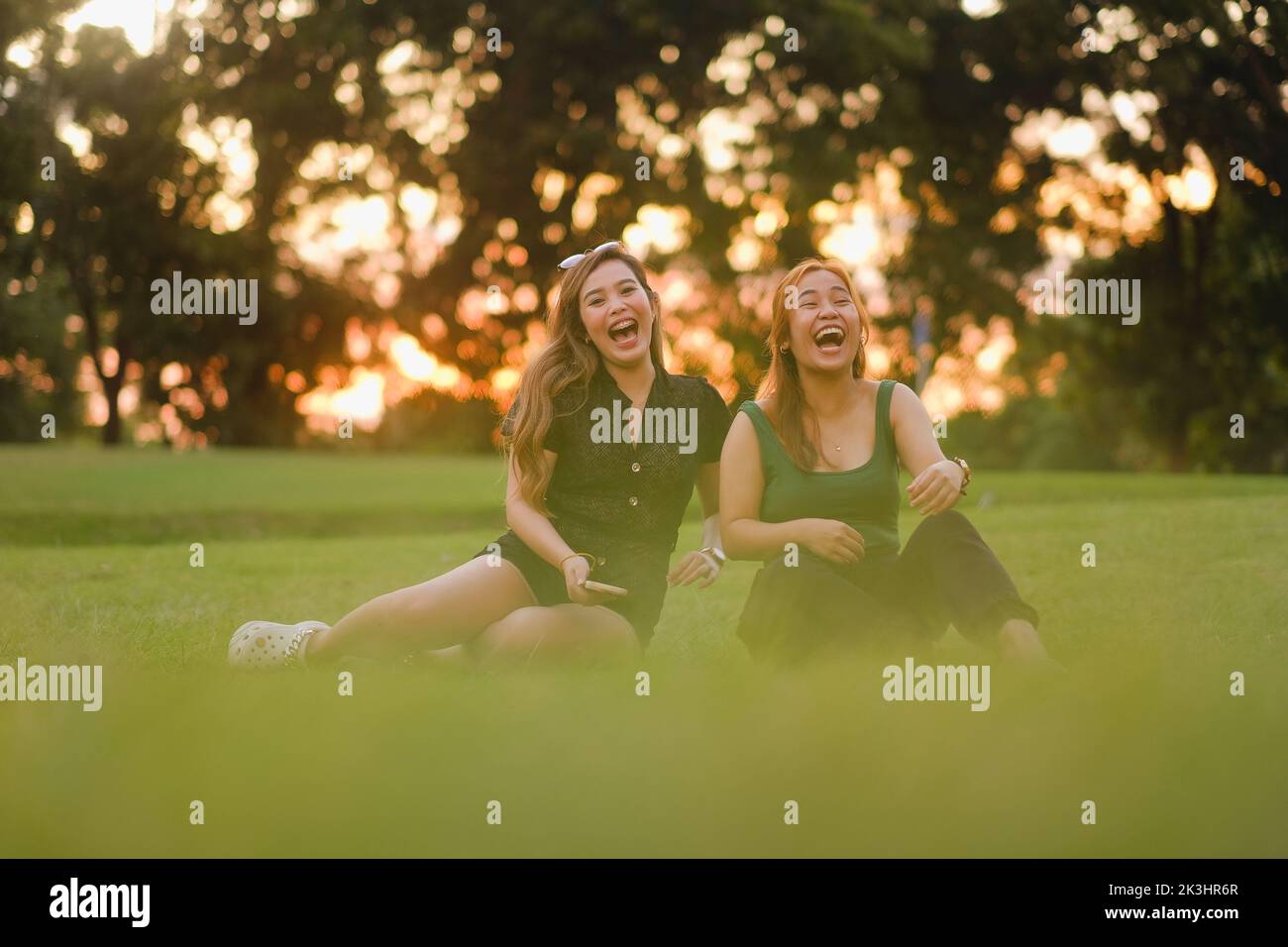 Two young Asian women, both with long wavy hair, howl with laughter. They are sitting beside each other on the grass on a beautiful summer day at sunset. Stock Photo