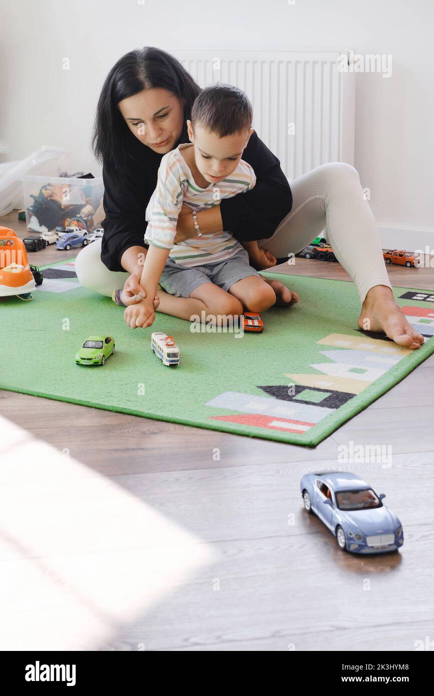 Disabled boy playing toy cars with mother at home. Cerebral palsy child entertaining on the mat with caregiver. Communication and rehabilitation in Stock Photo