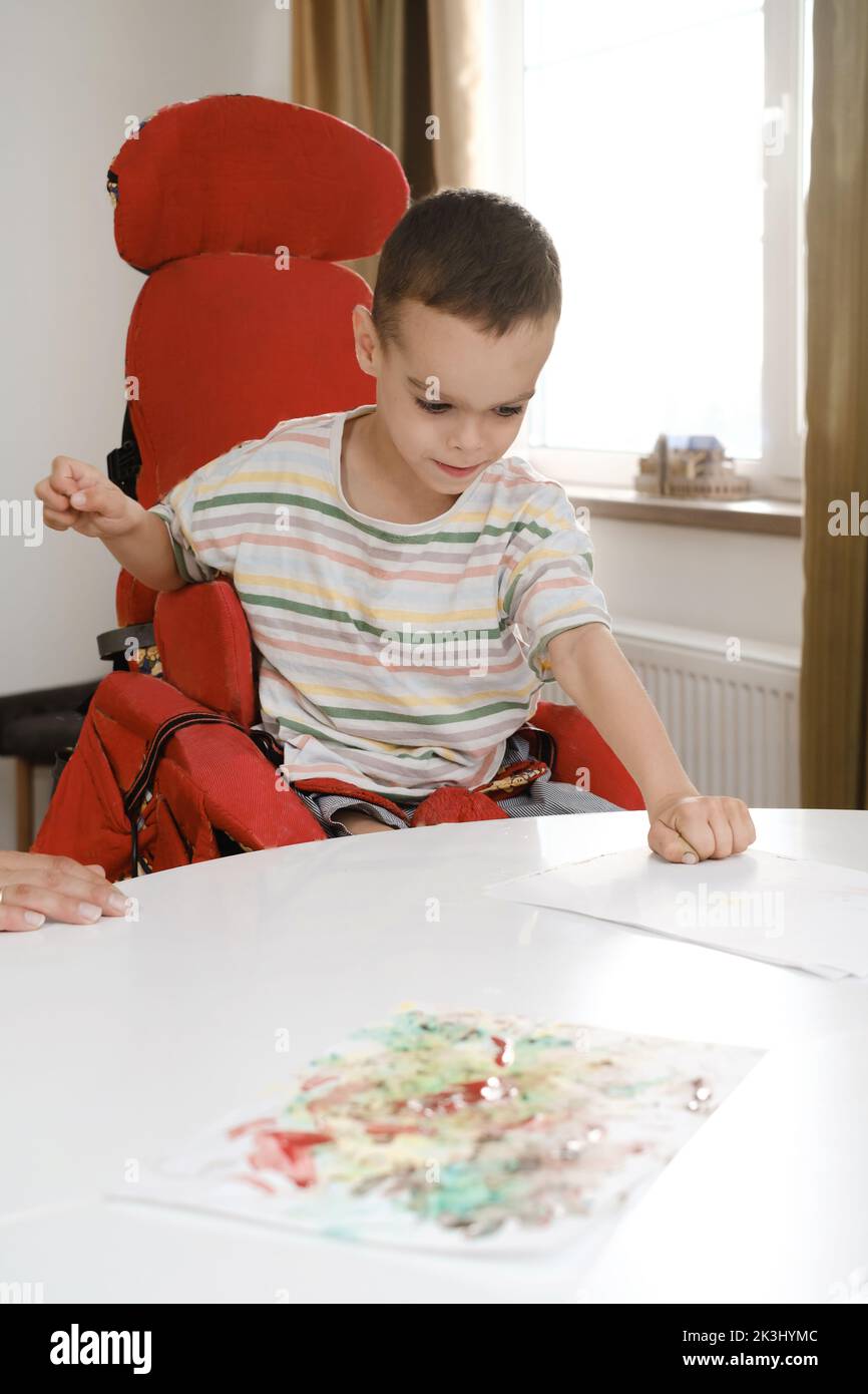 Child with cerebral palsy painting with fingers and hands. Happy handicap boy developing fine motor skills. Education for children with mental and Stock Photo