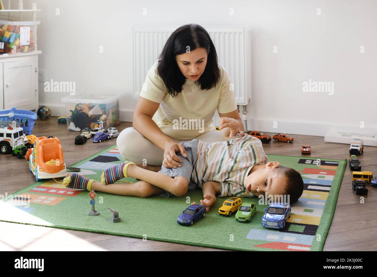 Disabled boy playing toy cars with mother at home. Cerebral palsy child entertaining on the mat with caregiver. Communication and rehabilitation in Stock Photo
