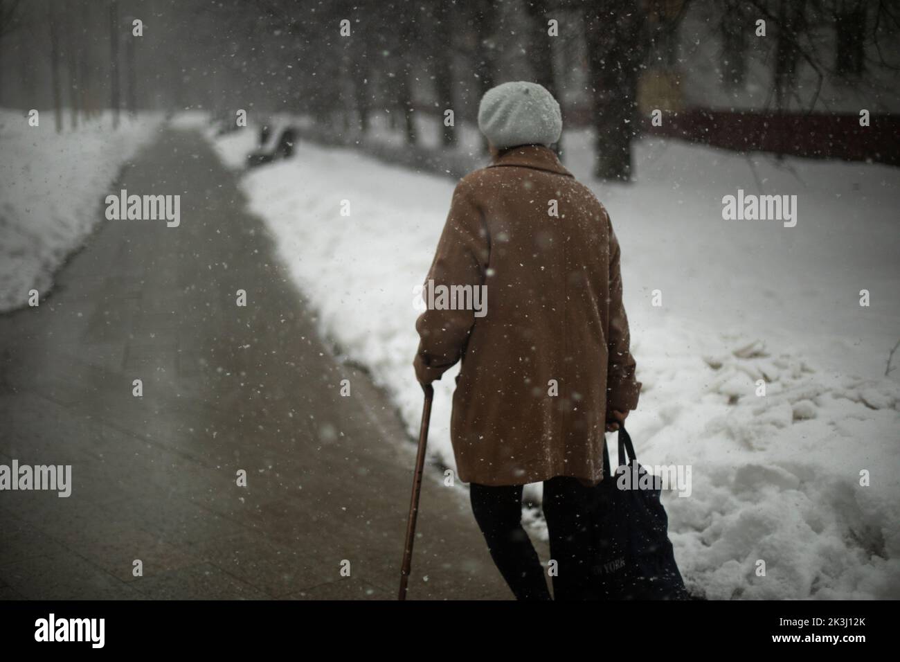 Woman walks in snow down street. Pensioner in winter in city. Woman leans on stick. Old man. Stock Photo