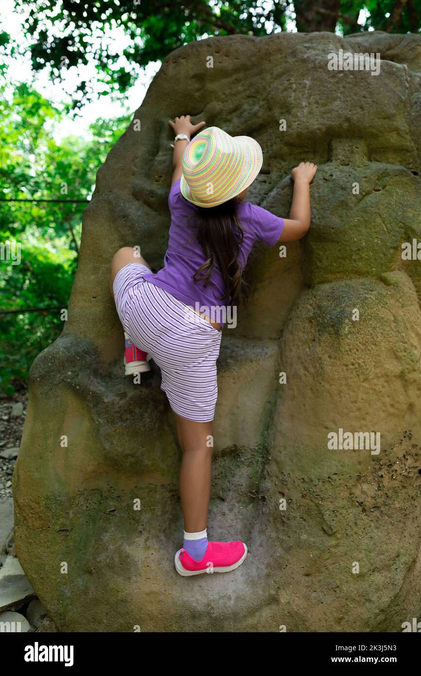Restless child in panama hat climb up rock in green forest back view. Little girl in comfortable clothes and footwear go down dangerous route. Tourism Stock Photo
