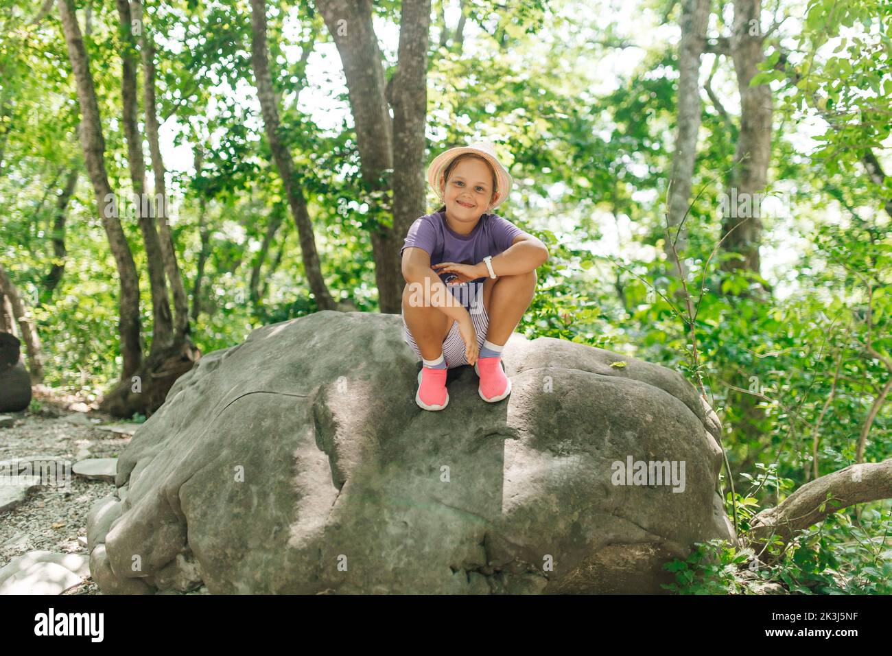 Happy smiling child in panama hat sit on big stone among growing trees on hill. Funny little girl walk and rest in forest on sunny day. Tourism Stock Photo