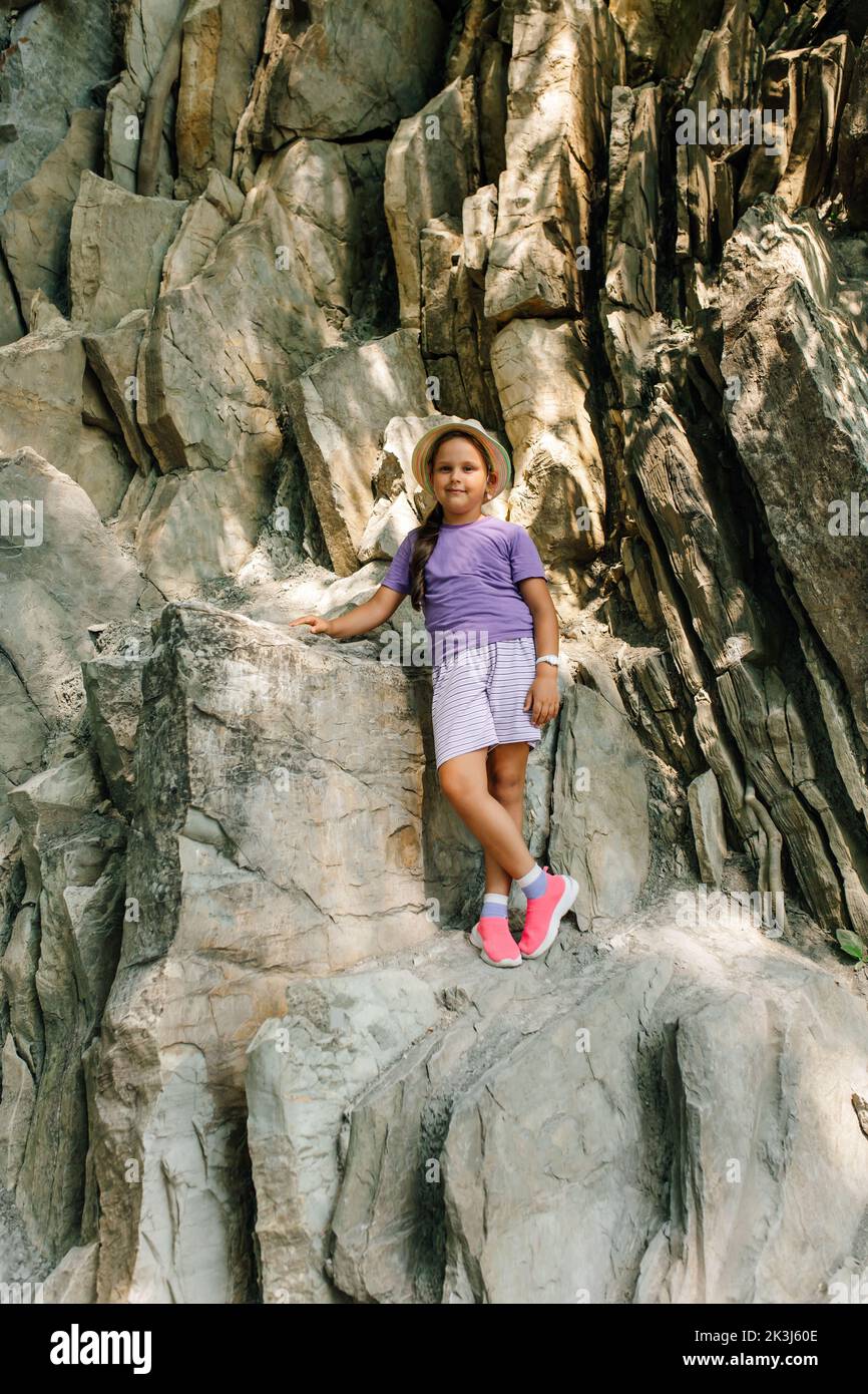 Nice child in panama hat stand leaning on big rock, mountain background. Little girl in sports clothing and sneakers walk and hike in mountains Stock Photo